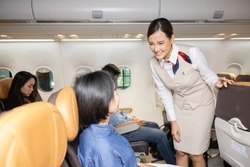 People in airplane cabin sitting and flying. A female flight attendant is speaking with a passenger sitting in the economy class making passengers comfortable throughout the journey.
