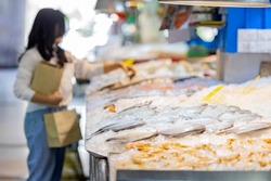 Fresh seafood on an ice counter in seafood zone at the supermarket. A counter with various seafood and Blurred woman holding brown paper bag, standing near seafood stall in background.  