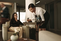 Business lounge waitress serving coffee to female passenger at waiting area. Business woman relaxing and waiting for flight at airport departure lounge.