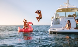 Man diving in the sea with friends sitting on yacht and inflatable toy. Group of friends enjoying a summer day on a inflatable toy and yacht.