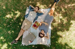 View from above of relaxed man and woman lying down at the park with hand basket, appetizing snacks and beers on blanket. Couple on picnic lying on blanket in grass outdoors.