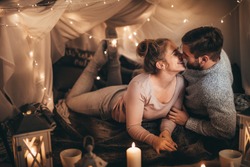 Couple together on bed in a room decorated with candle lights and tiny serial light bulbs. Smiling woman spending happy time with her husband in bedroom.