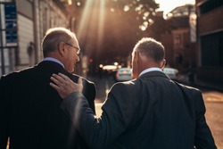 Rear view of two senior businessman walking together outdoors on city street. Mature businessman with hand in back of male colleague walking outdoors on a sunny day.