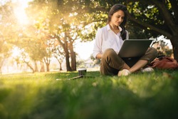 Woman sitting on grass at park working on laptop. Female wearing earphones using laptop while sitting under a tree at park with bright sunlight from behind.