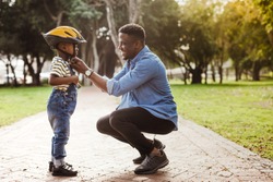 African man putting helmet on cute boy at the park. Father puts his son a protective helmet for riding bike.