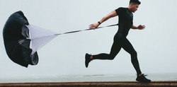 Strong man doing workout using resistance parachute outdoors. Young asian man in black sports clothing running with parachute.