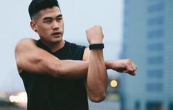 Fit young man with muscular build standing outside stretching hands and looking away. Asian fitness model doing warmup workout.