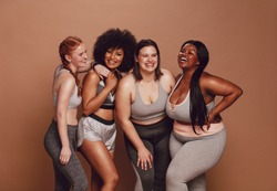 Smiling group of women in different size standing together in sportswear against brown background. Diverse group women looking at camera and laughing.