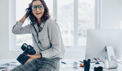 Smiling female photographer with a professional camera sitting on her desk. Woman with dslr camera in office looking away and smiling.