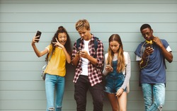 College friends standing outdoors looking using their cell phones. Young girl taking a selfie standing outdoors against a wall with her friends looking at their mobile phones.