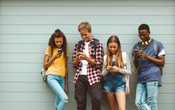 College friends wearing bags using mobile phones standing outdoors. Multiethnic teenage boys and girls standing outdoors against a wall and looking at their mobile phones.
