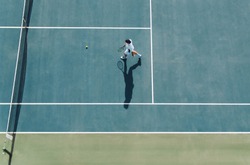 Aerial view of young male tennis player walking on hard court to pick the ball. Professional tennis player on club court.