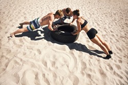 Small group of people doing push-ups on tire. Young athletes working out on beach during a hot summer day.