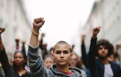 Young woman protesting on the street with her fist raised in air. Group of protesters on the road with their arms raised.