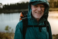 Portrait of a senior man carrying a backpack looking at camera and smiling. Fit old man on a hiking trip with river in background.