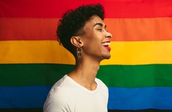 Smiling young gay man with make up standing against pride flag. Man with red lip stick and earring laughing in front of rainbow flag of gay pride.