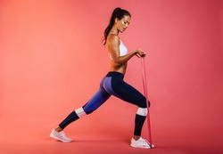 Slim athletic woman working on core strength training using resistance band. Side view of fit woman doing workout using stretch band on maroon background.