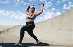 Full length of plus size woman exercising with resistance band. Female doing resistance band workout outdoors in morning.