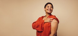 Woman showing her arm after receiving vaccine shot on brown background. Female with bandage on her arm looking away and smiling.