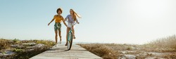 Excited woman riding bike down the boardwalk with her friends running along. Two female friends having a great time on their vacation. Panoramic shot with lots of copy space on background.