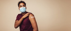 Man wearing face mask showing his vaccinated arm. Man in protective face mask received a corona vaccine looking away on brown background.