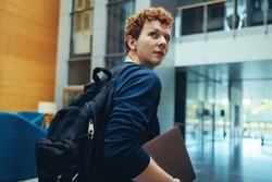College student carrying his bag and laptop in campus. Young man turning back over his shoulder and walking in college campus.