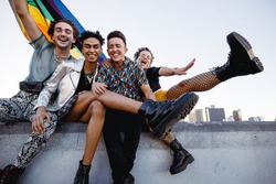 Four LGBTQ people celebrating pride while sitting together. Four friends smiling cheerfully while raising the rainbow pride flag. Group of young queer individuals celebrating together outdoors.