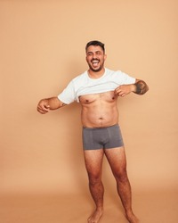 Flexing my natural body. Body positive young man lifting his shirt and smiling cheerfully while standing in a studio. Self-assured young man feeling comfortable in his natural body.