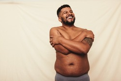 Man with pot belly embracing his natural body. Happy young man laughing cheerfully while standing shirtless against a studio background. Body positive man wearing underwear in a studio.