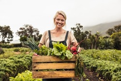 Cheerful organic farmer holding a box full of fresh produce on her farm. Happy young woman smiling at the camera while standing in her vegetable garden. Successful female farmer harvesting vegetables.