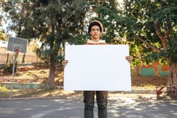 Teenage boy holding a blank placard in an urban park. Confident teenage activist displaying a white banner during the day. Teenager looking at the camera while standing alone outdoors.