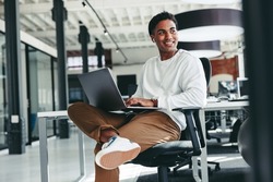 Cheerful software developer smiling in an office. Happy young businessman looking away while working on a laptop in a modern workplace. Creative businessman working on a new project.