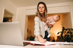 Design professional making notes while holding her baby. Multitasking mom planning a new project in her home office. Creative businesswoman balancing work and motherhood.