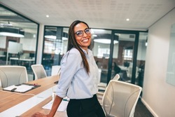 Mid-adult businesswoman smiling cheerfully in an office. Happy businesswoman leaning against a table in a boardroom. Cheerful female entrepreneur wearing business casual in a creative workplace.