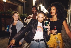 Group of happy friends dancing in the city at night. Four vibrant young people holding beer cans while walking together in the street. Friends having a good time on a weekend night.