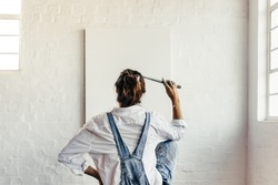 Contemplating creative ideas for a new project. Rearview of a young painter looking thoughtful while sitting in front of a blank canvas. Creative female artist holding a paintbrush to her head.