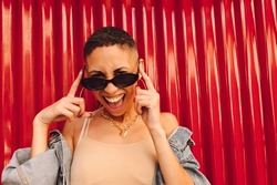 Cheerful young woman smiling at the camera while touching her sunglasses. Happy young woman standing against a red background. Young woman feeling vibrant and full of life.
