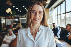 Successful young businesswoman smiling at the camera in a co-working space. Happy young female entrepreneur standing in a modern workplace with her co-workers in the background.