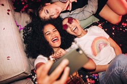 Vibrant selfies for vibrant people. Overhead view of three happy friends taking a selfie while lying on the floor at a house party. Group of cheerful female friends having fun together on the weekend.