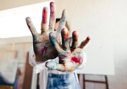 Painter blocking the camera from her face with her colour painted hands. Unrecognizable female artist standing in her art studio with her blank canvas in the background.