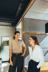 Successful businesswomen sharing a laugh in the office. Two young female entrepreneurs laughing cheerfully while standing together in a modern workspace.
