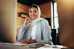 Happy Muslim businesswoman smiling at the camera while working on a computer in a modern office. Cheerful young businesswoman wearing a hijab in an inclusive workplace.