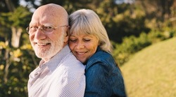 Happy mature couple embracing each other while standing in a park. Romantic elderly couple smiling and enjoying the sun together. Affectionate senior couple spending quality time after retirement.