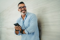 Mature entrepreneur smiling and looking away while standing against a wall in a modern workplace. Successful businessman sending a text message on his smartphone.