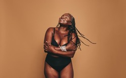 Loving my natural body. Happy woman with dreadlocks embracing her natural and ageing body. Mature black woman smiling cheerfully while standing against a studio background in black underwear.