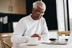 Senior businessman writing in a notebook while sitting at a desk in his home. Mature businessman making plans in his journal while doing remote freelance work.