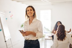 Happy young businesswoman smiling at the camera while holding a smartphone in a meeting room. Cheerful young businesswoman working in an all-female office.