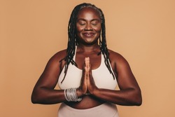 Mature woman meditating with her eyes closed and her hands in prayer position. Black woman with dreadlocks practicing yoga in a studio. Happy middle-aged woman maintaining a healthy lifestyle.