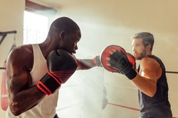 Athletic African man hitting the focus mitts held by his personal trainer in a gym. Young boxer having a training session with his coach.