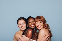 Three happy women with different skin tones smiling and embracing each other in a studio. Group of diverse women feeling comfortable in their natural skin. Body positive young women standing together.